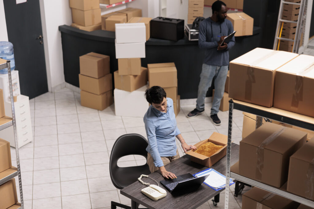 top view warehouse supervisor packing client clothes order cardboard box while checking shipping details laptop computer team working storehouse delivery department